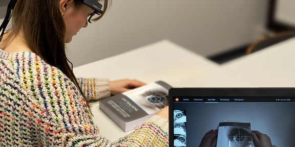 Eine Person nimmt an einem Eye-Tracking-Experiment teil und trägt mobile Tobii Pro Glasses 2. Die Daten des Experiments werden in Echtzeit auf einem Laptop angezeigt, während die Person ein Buch betrachtet.