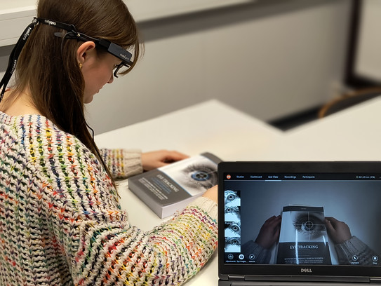 Eine Person nimmt an einem Eye-Tracking-Experiment teil und trägt mobile Tobii Pro Glasses 2. Die Daten des Experiments werden in Echtzeit auf einem Laptop angezeigt, während die Person ein Buch betrachtet.