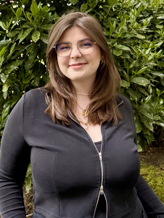 Elisa Die Tutorin Elisa steht vor einem Busch und lacht in die Kamera