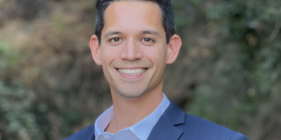 Portrait of PhD Wong: young adult, short black hair, smiling, standing in front of a blurred background of green bushes