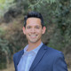 Portrait of PhD Wong: young adult, short black hair, smiling, standing in front of a blurred background of green bushes