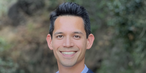 Portrait of PhD Wong: young adult, short black hair, smiling, standing in front of a blurred background of green bushes