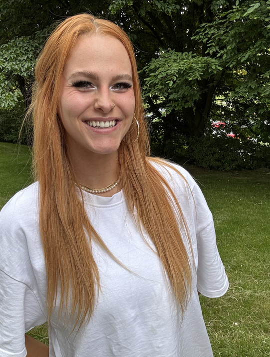 Foto von Madeleine. Sie steht vor einem Busch und lächelt in die Kamera