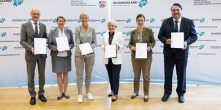 Vier Frauen und zwei Männer stehen mit Dokumenten in ihren Händen vor einer Stellwand mit der Aufschrift "Bildungsland NRW".