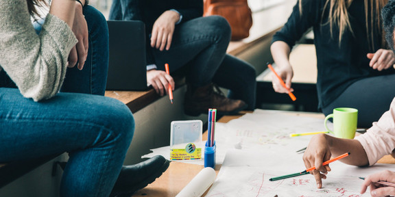 Brainstorming Vier Menschen sitzen an einem Tisch, die Gesichter sind nicht im Bild. Sie halten Stifte in der Hand und sehen nachdenkend aus. Eine Hand schreibt etwas auf das Plakat auf dem Tisch, welches sehr unstrukturiert gestaltet ist. Auf dem Tisch sind noch mehr Stifte und eine Tasse. Auf der Sitzbank erkennt man einen geöffneten Laptop und einen Rucksack im Hintergrund.