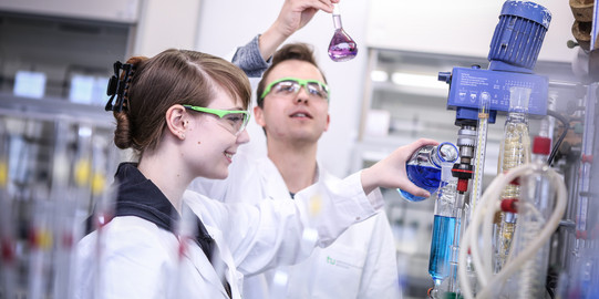 Zwei Auszubildende in einem chemischen Labor mit weißen Kitteln, einer gießt blaue Flüssigkeit in ein Destilliergerät.