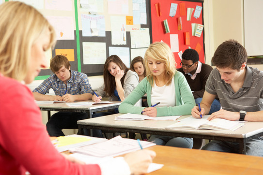 Schülerinnen und Schüler sitzen in einer Klasse und schreiben in Hefte, vor ihnen sitzt eine Lehrerin an einem Pult.