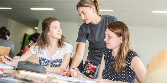 Zwei Personen sitzen mit Unterlagen und Stiften an einem Tisch. Eine dritte Person beugt sich zwischen die beiden Personen und spricht mit der Person, die links sitzt. Im Hintergrund sieht man weitere Personen an Tischen.