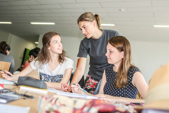 Zwei Personen sitzen mit Unterlagen und Stiften an einem Tisch. Eine dritte Person beugt sich zwischen die beiden Personen und spricht mit der Person, die links sitzt. Im Hintergrund sieht man weitere Personen an Tischen.