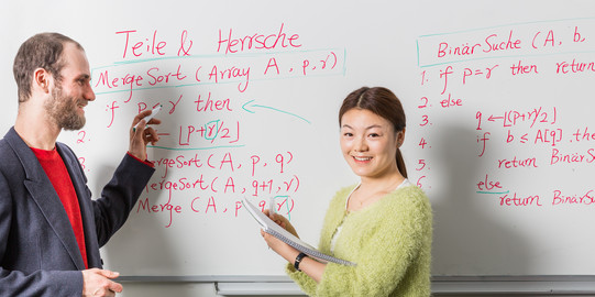Zwei Personen stehen vor einem Whiteboard mit Formeln und Beschriftung. Eine Person hat einen Stift in der Hand, die andere einen Block.