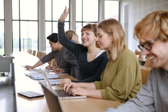 Fünf Studierende sitzen in einem Seminarraum an Tischen. Eine Studentin hebt ihre Hand.