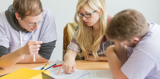 Drei Personen sitzen an einem Tisch und haben Unterlagen vor sich liegen. Die Person in der Mitte zeigt auf die Unterlagen und spricht.
