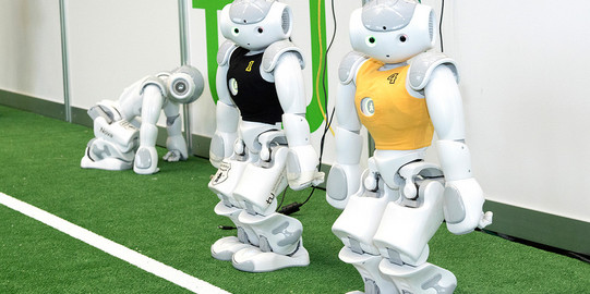 Zwei weiße Roboter im schwarzen und gelben Trikot stehen neben einander auf Kunstrasen, ein dritter Roboter kniet links hinter den beiden anderen Robotern.