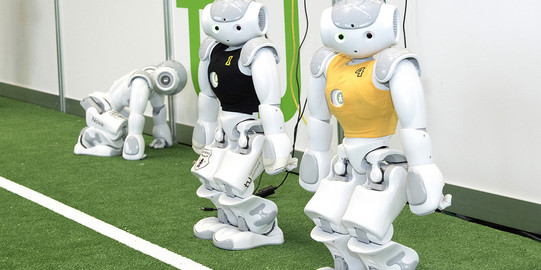 Zwei weiße Roboter im schwarzen und gelben Trikot stehen neben einander auf Kunstrasen, ein dritter Roboter kniet links hinter den beiden anderen Robotern.