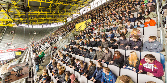 Erstsemesterbegrüßung Studierende des ersten Semesters sitzen bei der Erstsemesterbegrüßung auf den Rängen im Stadium und schauen seitlich einem Showact zu.